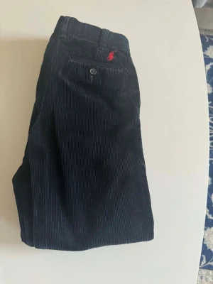 Mörkblå manchesterbyxor från Polo Ralph Lauren - Snygga mörkblå manchesterbyxor från Polo Ralph Lauren med klassisk rak passform. Byxorna har två bakfickor med knappar och det ikoniska röda Polo-logotypen broderad ovanför ena fickan. Tillverkade i mjuk manchester för en trendig look.