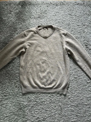 Ljusgrå tröja från Oscar Jacobson - Snygg beige tröja från Oscar Jacobson i klassisk modell med rund halsringning och långa ärmar. Tillverkad i mjukt merinoull som känns skönt mot huden. Perfekt att matcha med jeans eller chinos för en clean look. Tröjan är i mycket bra skick. Storlek M men passar S/XS då den har krympt, inga defekter.