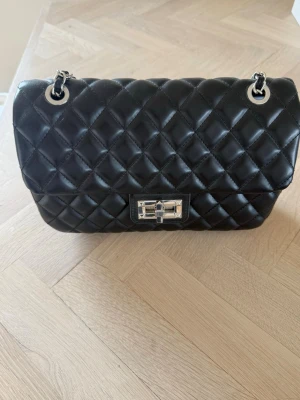 Svart quiltad axelväska med kedja - Säljer en svart quiltad axelväska med silverfärgad kedjerem och vridlås. Väskan har klassiskt rutmönster och stilren design, perfekt för dig som gillar tidlösa accessoarer. Materialet är glansigt konstläder och detaljerna är i metall.