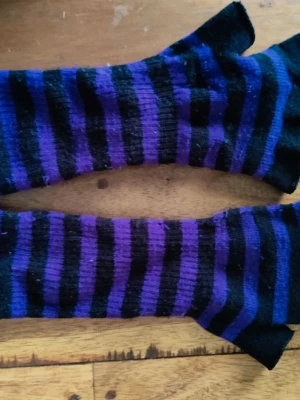 Lila och svarta randiga stickade vantar - Fingerlösa stickade vantar med randigt mönster i lila och svart. De har en mjuk och stretchig känsla och ger en alternativ vibe till din outfit. Perfekta för kyliga dagar när du vill ha fria fingrar.