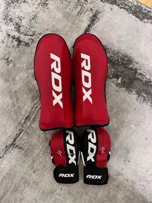 RDX handskar  - Säljer ett set med rödsvarta benskydd och handskar från RDX, perfekta för kampsport som MMA eller thaiboxning. Benskydden har bred kardborrestängning och handskarna har öppen tumme och justerbart handledsstöd. Snygg design med vit RDX-logga. Använda i kanske 2 månader. Ingen lukt då ja har tvättat de noggrant. Inga sår eller skador ( HANDSKARNA ÄR STORLEK: M & BENSKYDDEN ÄR STORLEK: S ) i