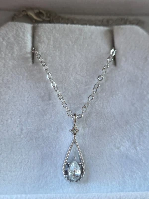 Silverfärgat halsband med droppformad kristall - Snyggt halsband i silverfärg med en tunn kedja och ett droppformat hänge. Hänget är dekorerat med små gnistrande stenar och en större klar kristall i mitten. Perfekt för dig som gillar stilrena och eleganta smycken. Materialet ser ut att vara metall med glittrande stenar.