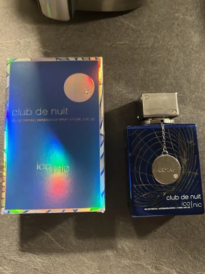 Armaf Club de Nuit Iconic parfym - Snygg parfym från Armaf, Club de Nuit Iconic, i en djupblå flaska med silverdetaljer och en metallbricka som hänger i kedja. Flaskan har en modern, fyrkantig form och kommer med en matchande holografisk blå förpackning. Eau de Parfum, 105 ml.