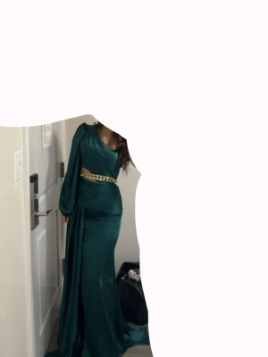 Grön långklänning med slits och kedjedetalj - Elegant långklänning i mörkgrönt med oneshoulder-design och lång ärm. Klänningen har en markerad midja med bred guldfärgad kedjedetalj och hög slits framtill. Materialet har en glansig finish som ger en lyxig känsla. Klänningen är endast använd en gång! Klänningen är i storlek 38 men den passar mer än 36!
