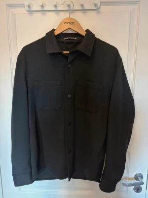 Svart overshirt från Five Units - Stilren svart overshirt från Zara med klassisk krage, knäppning framtill och två bröstfickor. Overshirten har raka ärmar och en enkel, clean design som passar till många olika looks. 