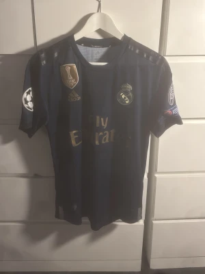 Real Madrid matchtröja Sergio Ramos - Mörkblå Real Madrid matchtröja från Adidas med guldiga detaljer och tryck. Tröjan har Champions League-märken på ärmarna och Sergio Ramos namn och nummer 4 på ryggen. Perfekt för fotbollsfans som vill visa sitt stöd för laget.