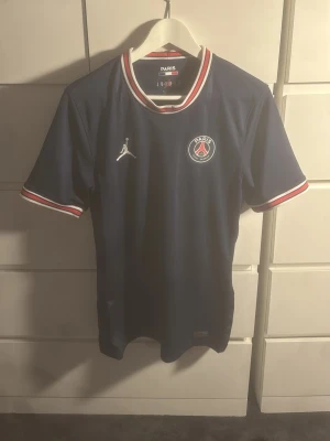 PSG matchtröja Sergio Ramos #4 - Mörkblå PSG matchtröja med vita och röda detaljer på krage och ärmslut. Framtill finns PSG-logga och Jordan-logga, på ryggen står det 'Sergio Ramos' och nummer 4. Perfekt för fotbollsfans som vill stötta sitt lag med stil.