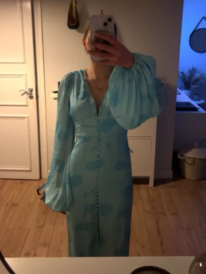 Adoore klänning  - Elegant ljusblå långklänning med blommigt mönster och v-ringning. Klänningen har långa, vida ärmar och klädda knappar framtill. Tillverkad i ett glansigt material som ger en lyxig känsla. Perfekt för speciella tillfällen. Den är från adoore i storlek 32, nypris 1695. Den har tappat en knapp längst ner men inget man tänker på💕