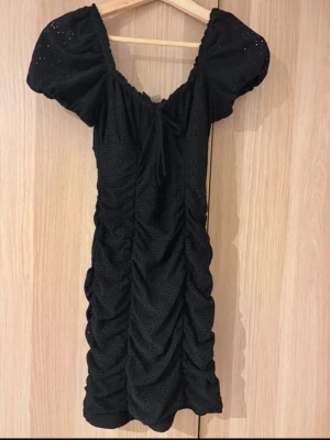 Svart kort klänning med puffärm - Snygg svart kort klänning med puffärmar och rynkade sidor. Klänningen har små hålmönster, knyt i ringningen och en feminin siluett. Perfekt för dig som vill ha en trendig och söt look.