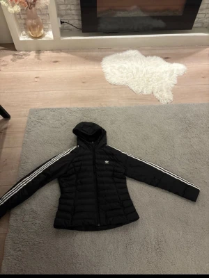 Svart pufferjacka från Adidas - Snygg svart pufferjacka från Adidas med huva och klassiska vita ränder längs ärmarna. Jackan har dragkedja framtill och Adidas logga på bröstet. Perfekt för kalla dagar.