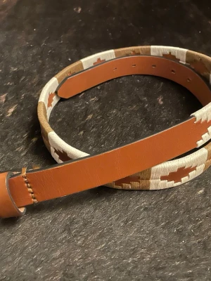 Brun och beige skinnbälte från Pampeano - Säljer ett snyggt bälte från Pampeano i brunt skinn med broderade detaljer i beige och vitt. Bältet har ett rutigt mönster och klassisk metallspänne. Perfekt accessoar för att lyfta din outfit med en unik touch. Använd 1 gång ska också sägas. Priset kan diskuteras