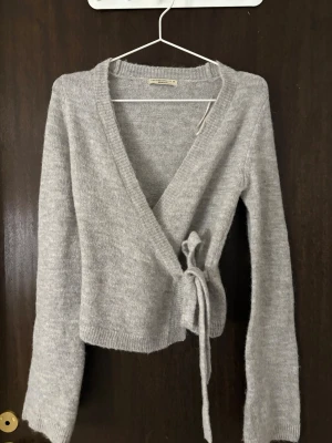 Grå omlottkofta från gina tricot - Mysig grå stickad omlottkofta från gina tricot med långa ärmar och knytband i midjan. Koftan har en djup v-ringning och ribbstickade muddar vid ärmslut och nederkant. Perfekt för lager på lager och enkel att styla. Helt nytt skick aldrig använd.