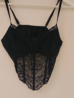 Svart spetstopp med tunna axelband - Säljer en snygg svart topp med tunna justerbara axelband och genomskinliga spetsdetaljer. Modellen har bygel och vackert blommönstrad spets framtill som ger en elegant look. Perfekt för dig som vill ha något extra till din outfit.