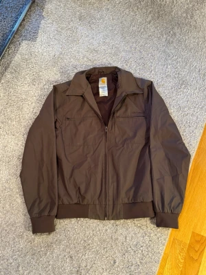 Carhartt Brun Bomberjacka - Säljer en brun Carhartt bomberjacka i storlek M.  Jackan är i fint skick utan slitage. Lätt och bekväm, perfekt för vår/höst eller svalare sommarkvällar.  Detaljer: 	•	Märke: Carhartt 	•	Storlek: M 	•	Färg: Brun 	•	Skick: Mycket bra 	•	Dragkedja framtill + fickor  Passar true to size. Hör av dig vid frågor eller för fler bilder!