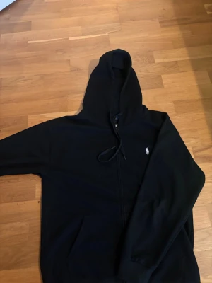 Svart hoodie från Ralph Lauren - Säljer en svart hoodie från Ralph Lauren med dragkedja framtill och klassisk huva med snören. Tröjan har det ikoniska vita Polo-logotypen broderad på bröstet och fickor framtill. Perfekt för en avslappnad och stilren look.