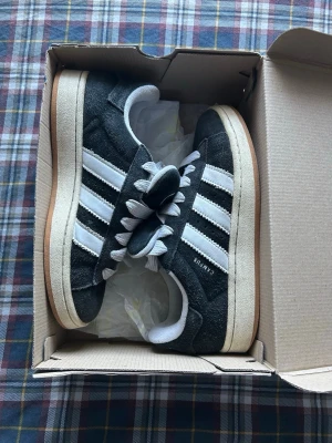 Svarta Adidas Campus sneakers - Säljer ett par svarta Adidas Campus sneakers med klassiska vita ränder och vit sula. Ovandelen är i mocka och skorna har en låg profil med gummisula i beige. Perfekta för dig som gillar retrostil och streetwear.