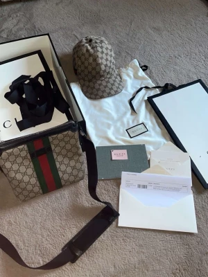 Gucci axelväska med GG-mönster - Säljer en snygg axelväska från Gucci i beige canvas med klassiskt GG-mönster och gröna/röda ränder framtill. Väskan har justerbar svart axelrem och svarta detaljer. Perfekt för dig som gillar lyxiga accessoarer och vill sticka ut.