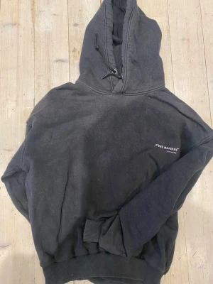 Svart hoodie från C'est Normal - Svart hoodie från C'est Normal med liten vit logga på bröstet. Tillverkad i en mjuk blandning av 47% ekologisk bomull, 41% bomull och 12% polyester. Klassisk huva med dragsko och ribbade muddar vid ärmar och nederkant. Perfekt för en chill och avslappnad stil.