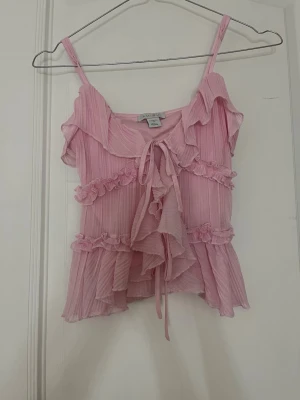 Rosa volang topp med knyt - Supersöt rosa topp från Urban Outfitters med tunna axelband, volangdetaljer och knyt framtill. Toppen är lätt och luftig med ribbad struktur och omlottkänsla. Perfekt för dig som gillar romantisk och trendig stil.