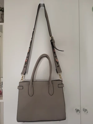 Beige handväska med blommig axelrem - Stilren beige handväska i fyrkantig modell med två handtag och en bred axelrem dekorerad med broderade blommor. Väskan har guldfärgade metalldetaljer och är tillverkad i slätt konstläder. Perfekt för dig som vill ha en trendig accessoar.