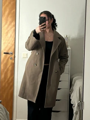 Beige dubbelknäppt kappa - Stilren beige kappa med dubbelknäppning och breda slag. Kappan har en rak passform och två sidofickor. Perfekt för dig som vill ha en klassisk och tidlös look.