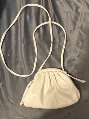 Vitl clutch väska  - Säljer en stilren vit handväska med lång axelrem och mjuk, rynkad design. Väskan har en unik formad öppning med metallspännen och är tillverkad i ett slätt, konstläderliknande material. Perfekt för dig som vill ha en enkel men trendig accessoar. Den är lite ben vit  alltså inte helt kritvit 
