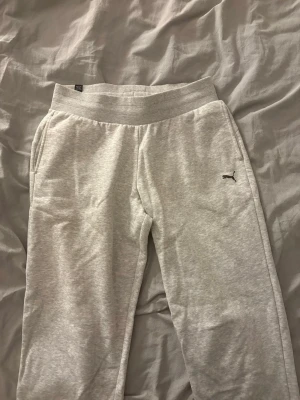 Ljusgrå mjukisbyxor från Puma - Ljusgrå mjukisbyxor från Puma med ribbad midja och diskret Puma-logga på höften. Byxorna har raka ben och sidofickor, perfekta för chill eller träning. Materialet är mjukt och bekvämt, troligtvis bomullsblandning.