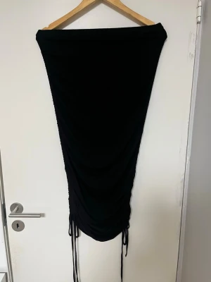 Svart långkjol med snörning - Trendig svart långkjol med snörning i sidorna som ger en snygg rynkad effekt. Kjolen har en smal passform och är perfekt för dig som gillar en stilren look med edge. Tillverkad i mjukt material som känns skönt mot huden.