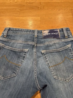 Blåa Jacob Cohën  jeans  - Säljer ett par klassiska blå jeans från Jacob Cohën med raka ben och snygga kontrastsömmar. Jeansen har två bakfickor och en tvättad finish som ger en avslappnad look. Perfekta för dig som gillar en tidlös stil.