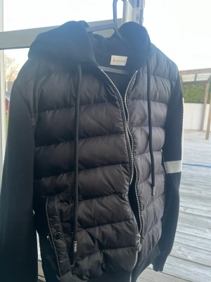 Svart cardiganjacka från Moncler - Svart cardiganjacka från Moncler med vadderad framsida i pufferstil och stickade ärmar. Jackan har huva med snörning, dragkedja framtill och en vit zigzag-detalj på ena ärmen samt Moncler-logga på axeln. Perfekt för en sportig och trendig look.