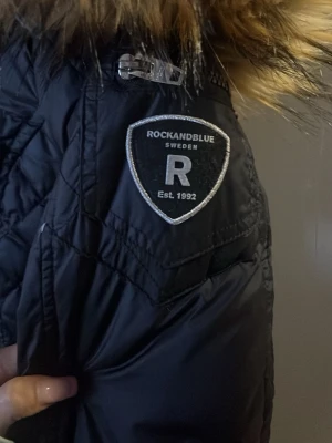 Svart dunjacka Rockandblue päls - Snygg svart lång dunjacka från Rockandblue med quiltade detaljer och avtagbar fuskpäls på huvan. Jackan köptes som ny för 3200 i butik 