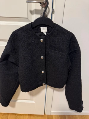 Svart bouclé cardigan med pärlknappar - Stilren svart cardiganjacka i bouclé-liknande material med raka axlar och croppad passform. Jackan har dekorativa pärlknappar framtill och vid ärmsluten samt en diskret bröstficka. Perfekt för att lyfta vilken outfit som helst.