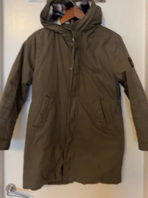 Olivgrön parkas från Replay - Säljer en olivgrön parkas från Replay med huva och rutigt innerfoder. Jackan har två snedställda fickor framtill och en patch med Replay-logga på ärmen. Lång modell som passar perfekt för kyliga dagar.