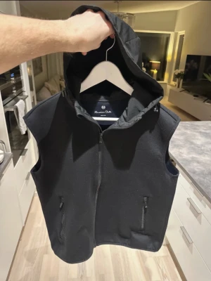 Svart dunväst med huva Massimo Dutti - Stilren svart dunväst från Massimo Dutti med dragkedja framtill och två fickor med dragkedja. Västen har en stor, skyddande huva och är tillverkad i ett mjukt, vadderat material som håller värmen. Perfekt för lager-på-lager och enkel att matcha.