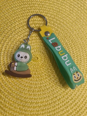 Labubu nyckelring med figur - Helt ny Söt nyckelring från Labubu med en grön och brun figur i djurdräkt och en matchande grön rem med texten 'Labubu' i färgglada bokstäver. Tillverkad i mjukt gummi och metall, perfekt att hänga på väskan eller nycklarna.  Se gärna mina andra annonser och skapa en bundle så får du rabatt. 