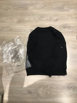 Svart sweatshirt från C.P. Company - Säljer en svart sweatshirt från C.P. Company med deras ikoniska linsdetalj på ärmen. Tröjan har rund halsringning och ribbade muddar vid ärmslut och nederkant. Tillverkad i mjukt bomullsmaterial som är skönt att bära. Perfekt för en clean och stilren look.