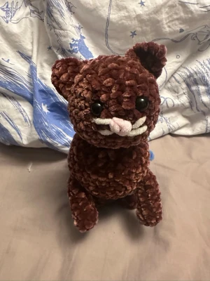 Brun virkad katt med blå kjol - Söt handgjord virkad katt i mjukt, fluffigt garn. Katten är brun med stora svarta ögon, vit nos och rosa morrhår. Den har en blå kjol virad runt kroppen. Som går att lätt ta av om man inte vill ha den där. Perfekt som dekoration eller gosedjur.