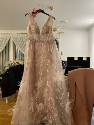 Champagnefärgad långklänning med spets - Elegant långklänning i champagnefärg med vackra spetsdetaljer, pärlbroderier och fjäderapplikationer. Klänningen har markerad midja och ett skirt, svepande tyg som ger en lyxig känsla. Perfekt för bal eller festliga tillfällen.