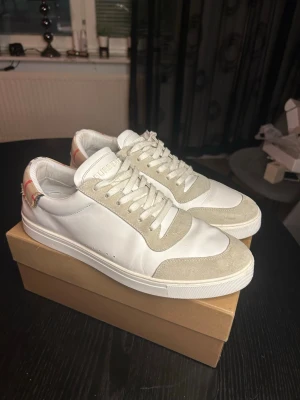 Burberry vita sneakers med beige mocka - Snygga vita sneakers från Burberry med beige mockadetaljer och klassiskt rutigt tyg på hälen. Skorna har vita snören, låg profil och diskret Burberry-logga bak. Perfekta för dig som gillar stilrena och exklusiva sneakers.