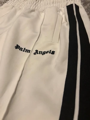 Vita mjukisbyxor Palm Angels - Säljer ett par vita mjukisbyxor från Palm Angels med svarta sidoränder och logga broderad på vänster sida. Byxorna har resår i midjan och raka ben. Materialet är mjukt och skönt, perfekt för en avslappnad stil. Storlek S