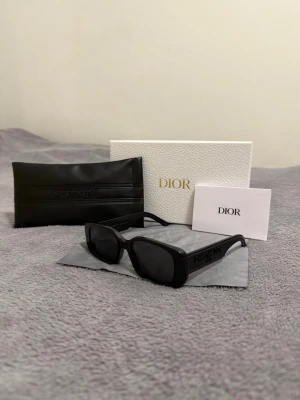 Svarta rektangulära solglasögon Dior - Snygga svarta solglasögon från Dior med rektangulära bågar och mörka glas. Klassisk och stilren design med Dior-logga på sidan. Kommer med originalfodral, putsduk och box. Perfekta för dig som vill ha en trendig accessoar.