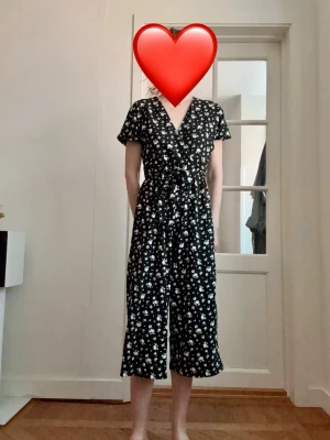 Svart vit blommig jumpsuit med knytband - Säljer en svart jumpsuit med vita småblommor och knytband i midjan. Modellen har korta ärmar, v-ringning och vida ben som slutar vid vaden. Perfekt för dig som gillar en avslappnad men ändå trendig stil. Stretchig och bekväm. Perfekt nu till våren och sommaren.