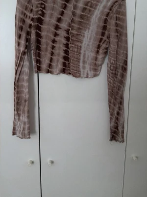 Brun och beige långärmad croppad topp - Trendig croppad topp med lång ärm i brunt och beige batikmönster. Toppen har vågiga sömmar framtill och är tillverkad i ett mjukt, stretchigt material. Perfekt för dig som gillar unika detaljer och en avslappnad stil.