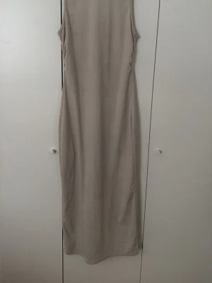 Beige långklänning ärmlös - Stilren beige långklänning med ärmlös design och rak passform. Perfekt för dig som gillar minimalistisk stil och vill ha en enkel men elegant look. Klänningen har rund halsringning och är tillverkad i ett mjukt material som faller snyggt.