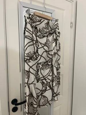 Vit omlottkjol med kedjemönster - Snygg lång omlottkjol i vitt med brunt kedjemönster. Kjolen har en knytning i midjan och volangdetaljer längs kanten. Skickas snabbt 💌
