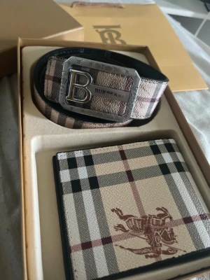 Rutig Burberry bälte med metallspänne - Säljer ett snyggt bälte från Burberry med klassiskt rutigt mönster i beige, svart och rött. Bältet har ett stort metallspänne med ett B och Burberry-logga. Perfekt accessoar för att lyfta din outfit. Storlek 105
