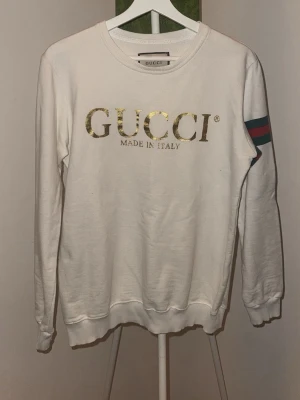 Vit sweatshirt  - Vit sweatshirt f med guldfärgat logotryck framtill och texten 'Made in Italy'. Tröjan har rund halsringning och klassiska gröna och röda ränder på ena ärmen. Mjuk bomullskänsla och avslappnad passform.