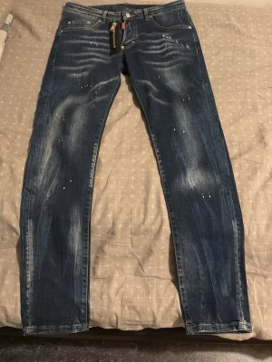 Blå jeans med färgstänk och slitningar - Säljer ett par blå jeans med coola färgstänk och slitna detaljer. Jeansen har klassisk femficksdesign, gylf med dragkedja och kontrastsömmar. Perfekta för dig som gillar en avslappnad och trendig stil.
