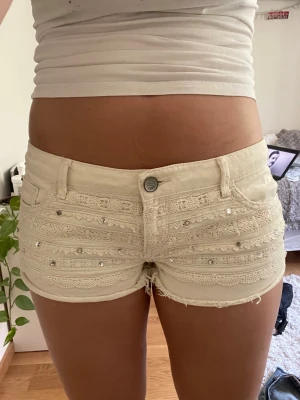 Beiga spets- och pärlshorts - Snygga beiga shorts med spetsdetaljer och små pärlor framtill. Shortsen har låg midja, råa kanter och klassisk femficksdesign. Perfekta för sommaren och ger en söt och trendig look.
