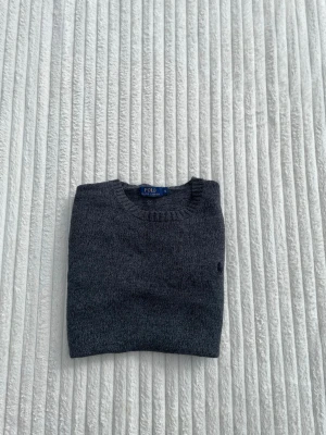 Mörkgrå tröja från Polo Ralph Lauren - Mörkgrå stickad tröja från Polo Ralph Lauren med rund halsringning och diskret broderad logga på bröstet. Tröjan har ribbade muddar vid ärmslut och nederkant. Perfekt för en stilren och klassisk look.